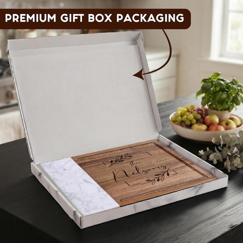 May include: A square wooden cutting board with a white marble accent piece, presented in an open white box with marble-patterned exterior sides. The board features the engraved text "The Nelsons" surrounded by a decorative leaf frame design.