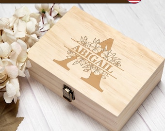 May include: A rectangular light-coloured wooden box with a metal latch. The lid features a laser-engraved design of a large letter A intertwined with floral illustrations and the name ABIGAIL. A banner at the top reads LASER ENGRAVED IN USA.