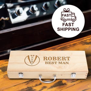 May include: A light-colored wooden box with a silver handle and clasps. The box is engraved with a tuxedo graphic, the name "ROBERT", and the text "BEST MAN". The box is on a wooden surface.