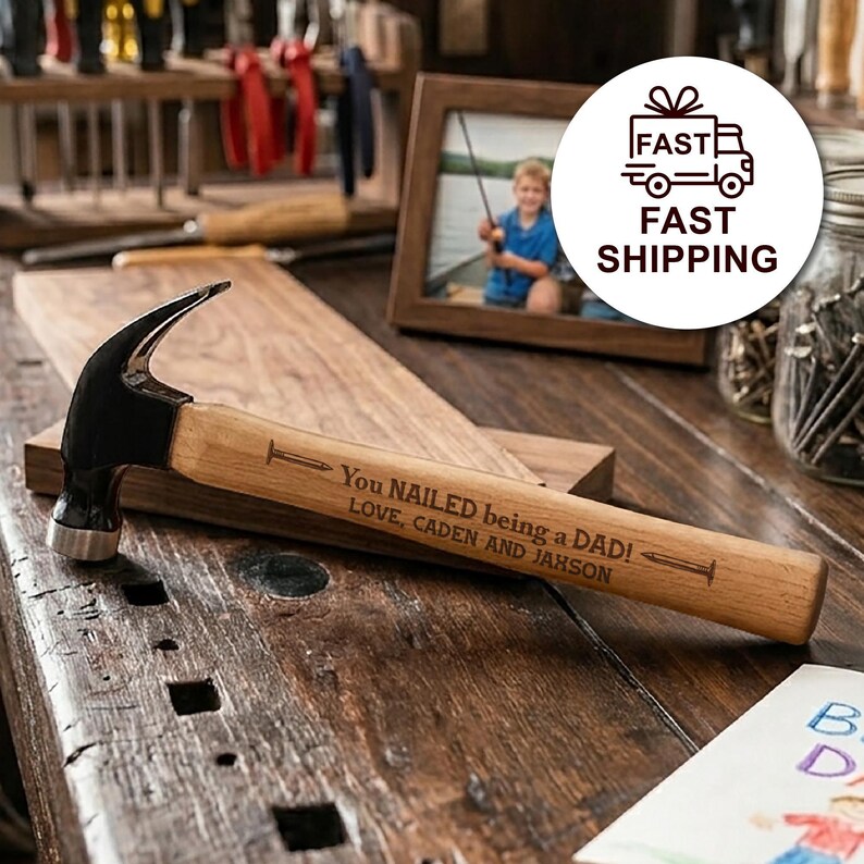May include: A claw hammer with a black metal head and a light brown wooden handle. The handle is engraved with the text: You NAILED being a DAD! LOVE, CADEN AND JAHSON, featuring small nail illustrations on either side of the main message.