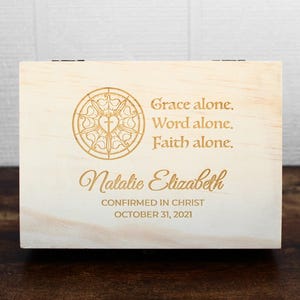 K&ouml;nnte beinhalten: Eine hellfarbene Holzkiste mit Scharnierdeckel. Die Kiste hat eingravierten Text und ein dekoratives Emblem. Der Text lautet "Grace alone. Word alone. Faith alone." und "Natalie Elizabeth CONFIRMED IN CHRIST OCTOBER 31, 2021."