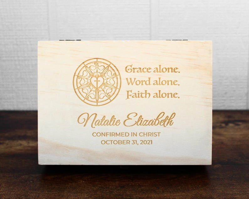 K&ouml;nnte beinhalten: Eine hellfarbene Holzkiste mit Scharnierdeckel. Die Kiste hat eingravierten Text und ein dekoratives Emblem. Der Text lautet "Grace alone. Word alone. Faith alone." und "Natalie Elizabeth CONFIRMED IN CHRIST OCTOBER 31, 2021."