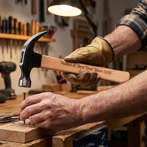 May include: A claw hammer with a light-colored wooden handle featuring the engraved text 'Nailed it! Best Dad Since 2026'. The hammer head is made of dark metal, and the handle is held by a tan leather work glove while driving a metal nail into wood.