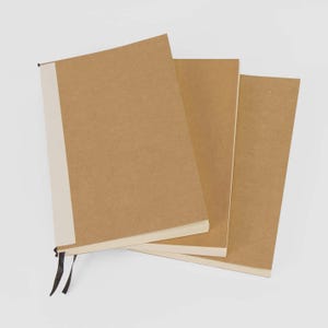 May include: Three brown notebooks with cream-colored pages and a light beige spine. The notebook on the left has a black ribbon bookmark. The notebooks are stacked slightly, showing the edges of the pages.