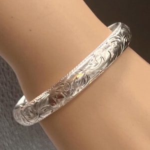 Może przedstawiać: Srebrzysta bransoletka typu bangle z misternym, grawerowanym wzorem kwiatowym na całej zewnętrznej powierzchni. Metalowa obręcz ma polerowane, odblaskowe wykończenie ze szczegółowymi, wgłębionymi liniami, które tworzą teksturowany wygląd.