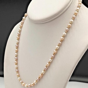 Puede incluir: Un delicado collar con pequeñas perlas redondas alternadas con cuentas doradas. Las perlas son blancas y rosa pálido. El collar se exhibe en un expositor de joyas blanco sobre un fondo oscuro.