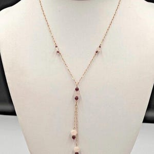 Puede incluir: Un delicado collar de cadena dorada con un diseño en forma de Y. La cadena está decorada con pequeñas cuentas esféricas de color rosa pálido y rojo oscuro espaciadas a lo largo, terminando en una borla colgante de cuentas en el centro.