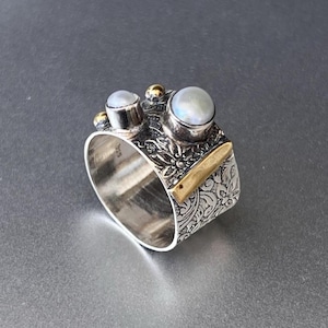 Puede incluir: Un anillo de plata con un diseño floral y detalles dorados. El anillo presenta tres piedras redondas e iridiscentes. La banda tiene un patrón floral grabado y un acento rectangular dorado.