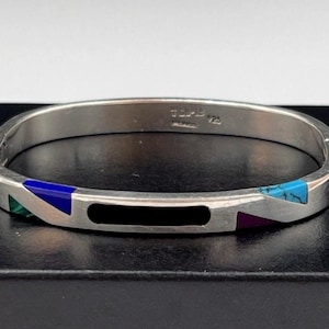 May include: A silver hinged bangle bracelet featuring inlaid geometric segments of black, blue, purple, green, and turquoise stone. The interior of the band is stamped with the text TLP-B and 925, indicating sterling silver composition.