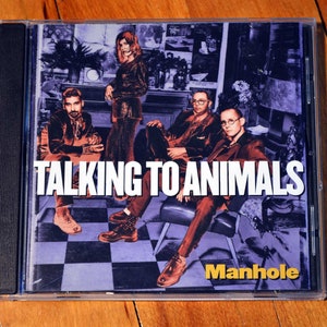 Puede incluir: Una caja de CD con una columna vertebral negra y una imagen colorida de una banda llamada "Manhole" en la parte delantera. La banda se llama "Talking to Animals" y la portada del álbum presenta a cuatro personas en una habitación a cuadros.