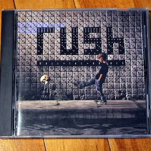 May include: A CD case with a black spine and a clear front cover. The front cover features a black and white image of a person kicking a skull against a wall with a tiled pattern. The wall has the word "QUEER" written in large letters. The text "ROLL INTO THE NIGHT" is written below the word "QUEER".