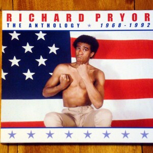 May include: A CD case featuring Richard Pryor. The cover shows Pryor in a fighting stance against an American flag backdrop. The title "RICHARD PRYOR THE ANTHOLOGY 1968-1992" is printed in red and white.