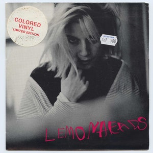 May include: A black and white photo of a woman with short blonde hair. The photo is a record cover for the band Lemonheads. The text "Colored Vinyl Limited Edition" is on a white circle sticker in the top left corner. The text "Mystery Train 727/500" is on a blue sticker in the top right corner.
