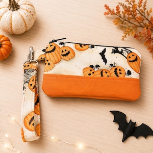 May include: A rectangular fabric wristlet pouch featuring a cream-colored background with a pattern of orange jack-o'-lanterns and black bats. The bottom section is solid orange fabric, and it includes a matching wrist strap with a metal clasp.