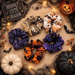 May include: Five fabric hair scrunchies featuring Halloween-themed patterns including pumpkins, spider webs, and autumn leaves. The scrunchies are arranged in a circular formation alongside a sign that reads SPOOKY CUTE SEASON.
