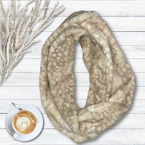 May include: A beige infinity scarf with a spotted pattern. The soft, faux fur scarf is draped on a white wooden surface. A cup of coffee with latte art and decorative branches are also visible.