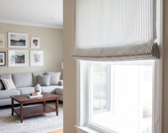 May include: A white window shade with vertical black stripes is partially lowered, covering a window. The shade is mounted inside a white window frame. The room has a neutral color palette with a gray sofa and framed artwork.