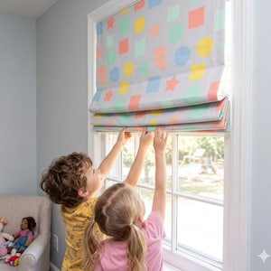 May include: A gray Roman shade with colorful geometric shapes, including squares, circles, and stars, is partially lowered. The window treatment is being adjusted by two children. The room has light blue walls and a neutral-toned chair.