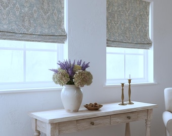 May include: Two windows featuring light blue and beige paisley patterned roman shades. A white ceramic vase filled with purple lavender and green hydrangea sits on a light wood console table with two drawers, alongside a small bowl and two candlesticks.