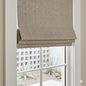 Puede incluir: Una persiana romana de tono neutro está parcialmente bajada, mostrando su tejido texturizado. La persiana está instalada dentro de un marco de ventana blanco, ofreciendo una vista del exterior. La ventana presenta un patrón de rejilla y vegetación.