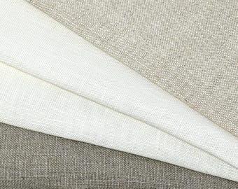May include: Close-up of folded linen fabric samples in shades of white and gray. The fabric has a textured weave, with the samples layered to show the different colors and textures. The image highlights the natural look of the linen.