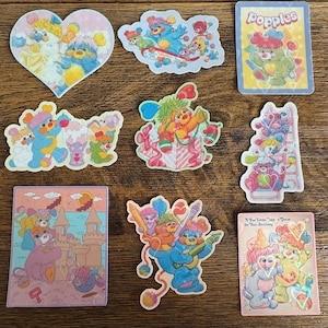 May include: A collection of colorful vintage Popples stickers. The stickers feature cartoon characters in various poses, including a heart-shaped sticker with two Popples. Other stickers show Popples with ice cream, on a cloud, and in a gift box.