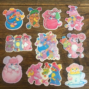 May include: A set of 11 colorful cartoon character stickers. The stickers feature various characters with pink, blue, yellow, and white colors. The characters are all smiling and have different expressions. The stickers are perfect for adding a touch of whimsy to any project.