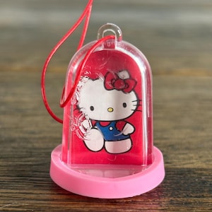 May include: A clear plastic bell-shaped ornament featuring Hello Kitty. The ornament has a red background with a Hello Kitty graphic, a pink base, and a red string for hanging. The ornament is approximately 2 inches tall.