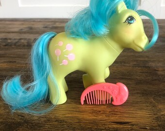 May include: A light green My Little Pony with a blue mane and tail. The pony has pink and white lollipop-shaped markings on its side. The pony is holding a pink comb with a star design.