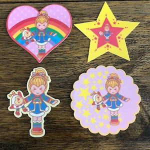 May include: Four colorful iron-on patches featuring a rainbow, stars, and cartoon characters. The characters are wearing colorful outfits and holding small animal companions. The patches are perfect for adding a touch of whimsy to clothing, bags, or other fabric items.