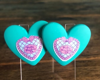 May include: Two teal heart-shaped earrings with a pink and purple Polly Pocket logo on each.