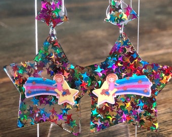 May include: A pair of star-shaped earrings with a rainbow and a girl riding a shooting star design. The earrings are filled with iridescent glitter.