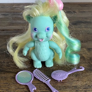 May include: A mint green dragon figurine with long, pale yellow hair and a pink hair clip. The dragon has large purple eyes and a textured chest. Included are three purple plastic grooming accessories: a round hand mirror, a comb, and a hairbrush.