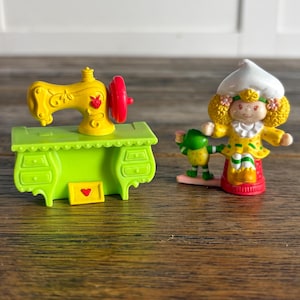 May include: A yellow toy sewing machine with a red wheel sits on a green table. Next to it is a figurine of a girl in a yellow dress and white hat, holding a green creature. The toys are on a wooden surface.