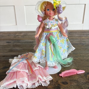Op de afbeelding: Een pop met lang, meerkleurig haar draagt een bloemenjurk, een witte hoed met kanten rand en gele bloemen, en een paars accessoire. Naast de pop ligt een aparte roze gelaagde jurk met kanten rand en een kleine roze plastic haarkam.