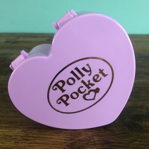 Puede incluir: Un juguete Polly Pocket de color morado en forma de corazón con un logotipo dorado que dice "Polly Pocket" con un pequeño símbolo de corazón.