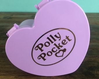 May include: A purple heart-shaped Polly Pocket toy with a gold logo that reads "Polly Pocket" with a small heart symbol.