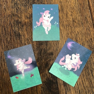 May include: Three cards featuring a pink and white unicorn wearing a pink tutu and dancing on a green background with stars and a crescent moon.