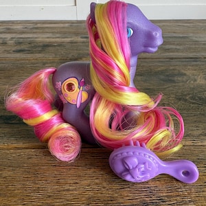 May include: A purple toy pony with a colorful mane and tail in shades of pink, yellow, and orange. The pony has a painted design on its flank and a matching purple brush. The toy is designed for children.