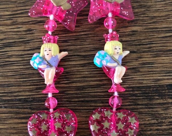 May include: A pair of pink dangle earrings with a star, a heart, a small figurine of a girl, and another heart with gold stars.