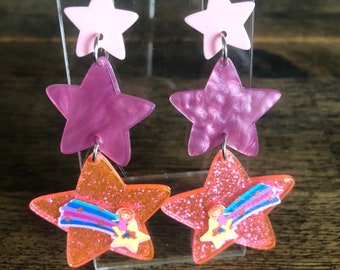 May include: A pair of dangle earrings with a pink, purple, and orange star design. The stars are layered on top of each other and feature a glitter finish. The bottom star has a small cartoon image of a girl riding a shooting star.