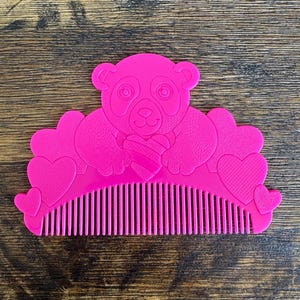 Puede incluir: Un peine rosa brillante con un diseño de oso panda sosteniendo un corazón. El peine tiene una fila de dientes y detalles en forma de corazón. El oso panda tiene una cara sonriente y sostiene un corazón a rayas.