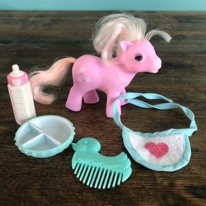 Puede incluir: Un pony bebé rosa con una melena y cola blancas, un biberon azul, un plato dividido azul y blanco, un babero azul y blanco con un corazón rosa y un peine azul claro con forma de pato.