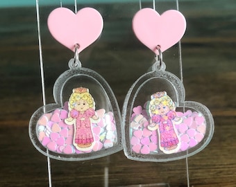 May include: A pair of pink heart-shaped earrings with clear resin hearts containing glitter and a princess doll image.