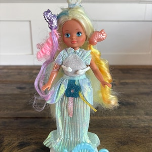 May include: A doll with long, multi-colored hair featuring pink, yellow, and blue strands. The doll wears a shimmering, iridescent light blue gown with a silver bodice and a dark blue flower accent. A light blue plastic comb is positioned at the base.