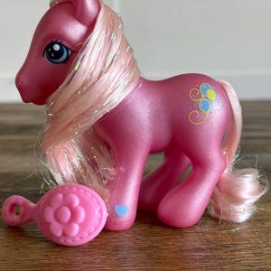 May include: A pink toy pony with a flowing, glittery mane and tail. The pony has blue eyes and a decorative design on its flank. A pink flower-shaped brush is next to the pony. A blue heart is on the pony's leg.