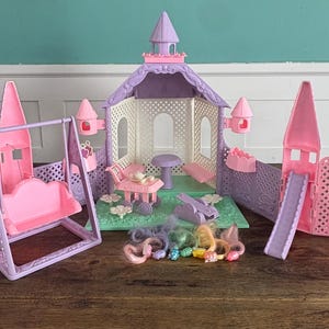 Könnte beinhalten: Ein Vintage-Spielset in Rosa- und Lilatönen mit einem Schloss, einer Schaukel und einer Rutsche. Das Set enthält Miniaturmöbel und bunte Haaraccessoires, die eine skurrile Szene schaffen.