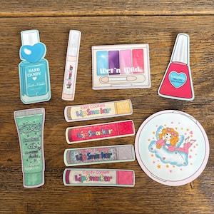 Puede incluir: Una colección de pegatinas coloridas con productos cosméticos y un ángel de dibujos animados. Incluye un frasco de esmalte de uñas, brillo labial, una paleta de sombras de ojos y tubos de crema. También hay pegatinas de bálsamo labial Lip Smacker.