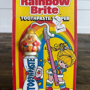 May include: Rainbow Brite Toothpaste Topper package. The packaging is yellow with a rainbow design and features a cartoon girl holding a pink toothbrush. The toothpaste tube has a cartoon character on it. The text reads "Makes Brushing Fun!!!"