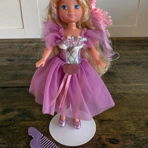 Könnte beinhalten: Eine Puppe mit blondem Haar, das rosa und violette Strähnen aufweist, trägt ein violettes Tüllkleid mit silbernem Oberteil und einer rosa Blumenapplikation. Die Puppe steht auf einem weißen runden Sockel und hat einen violetten Kamm.